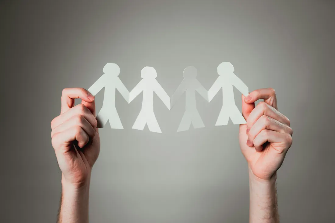 Two hands holding up a chain of paper cut-out people, with one figure faded out of the line.